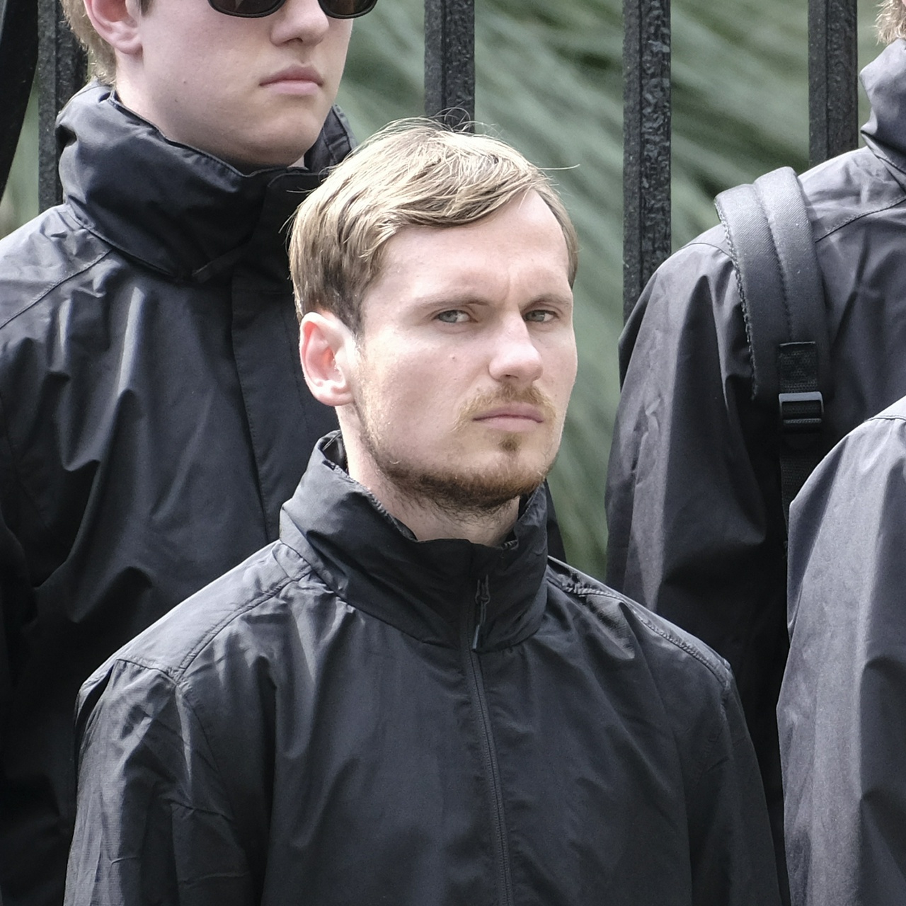 A short man with greased brown hair sneers worriedly at the camera. He has facial hair that travels a fair way down his neck, and a mole or pimple under his right eye