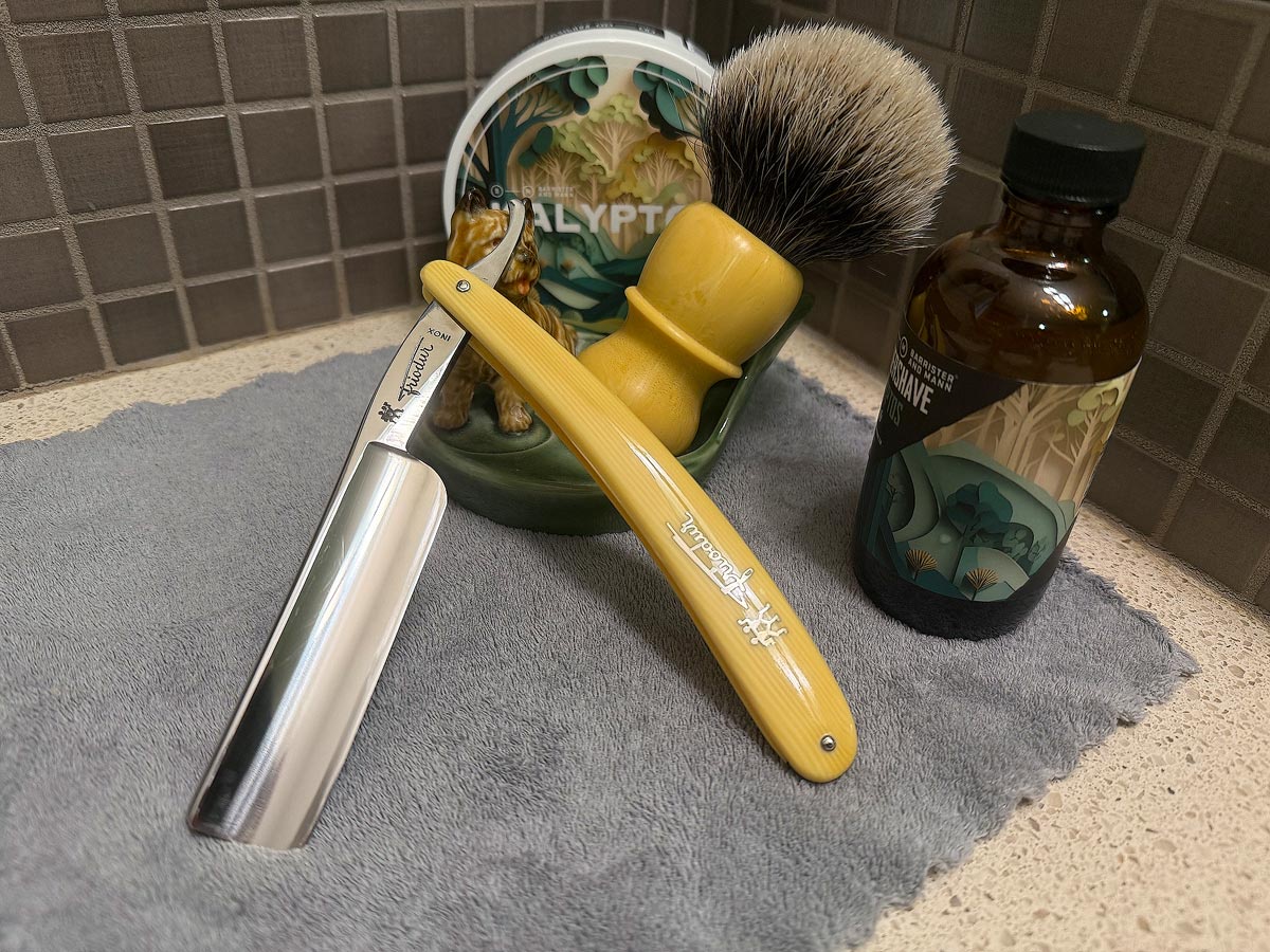On a gray microfiber towel a straight razor with blade open about 45 degrees leans next to a bottle of aftershave and a jar of shave soap. A shave brush rests bristles-up in a green Wade ceramic pipe rest that features a brown Cairn Terrier.