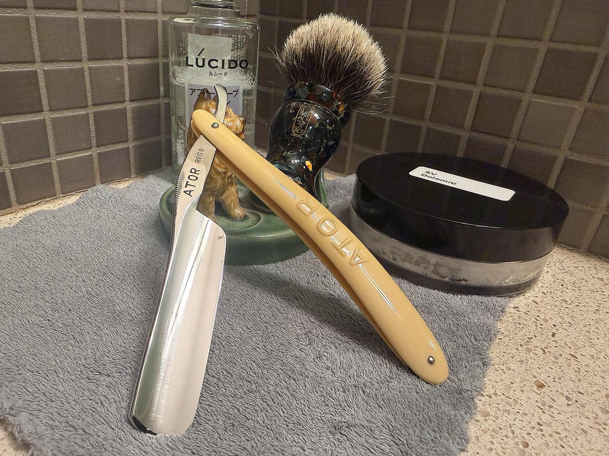 On a gray microfiber towel a straight razor with blade open about 45 degrees leans next to a bottle of aftershave and a jar of shave soap. A shave brush rests bristles-up in a green Wade ceramic pipe rest that features a brown Cairn Terrier.