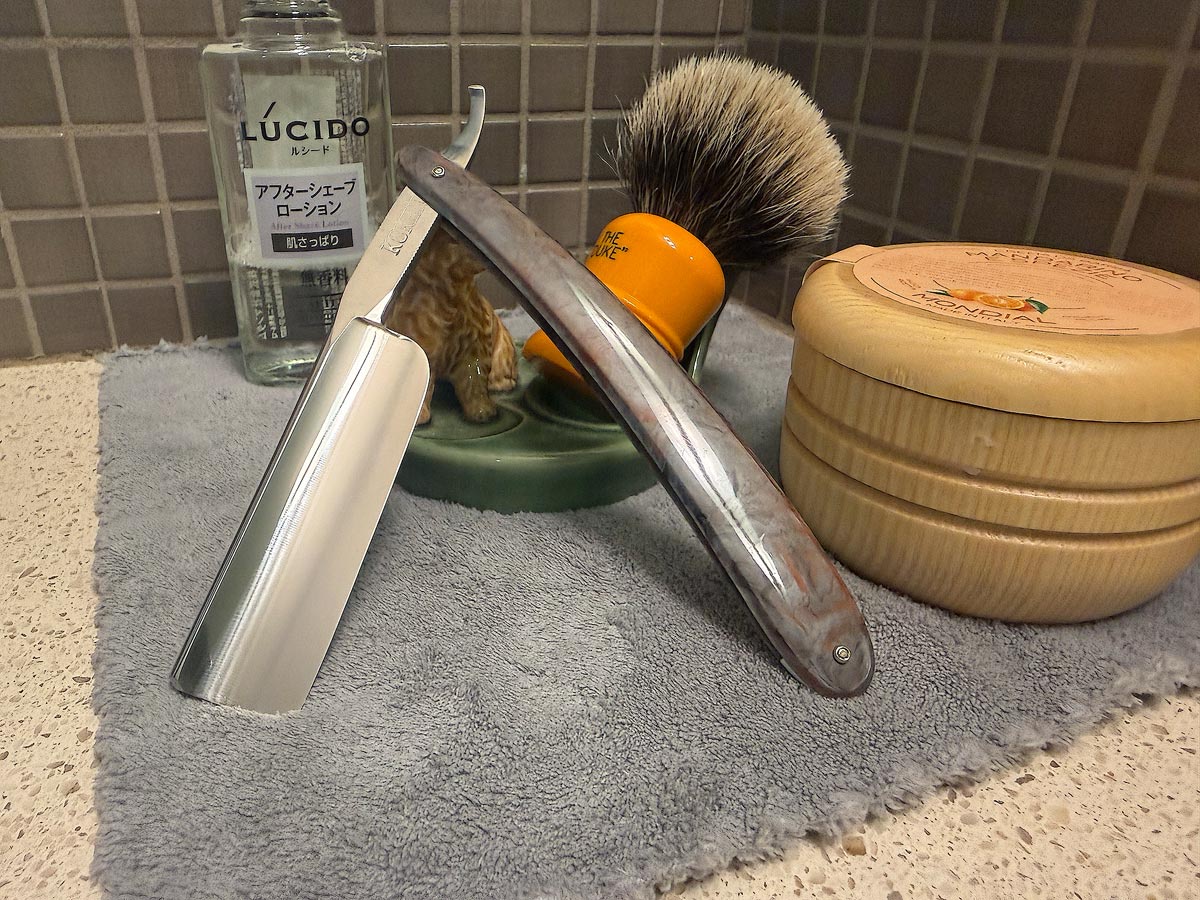 On a gray microfiber towel a straight razor with blade open about 45 degrees leans next to a bottle of aftershave and a jar of shave soap. A shave brush rests bristles-up in a green Wade ceramic pipe rest that features a brown Cairn Terrier.