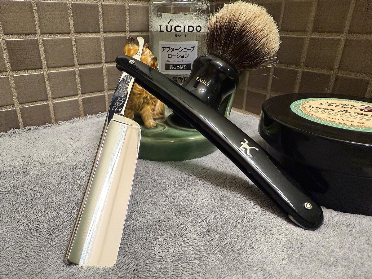 On a gray microfiber towel a straight razor with blade open about 45 degrees leans next to a bottle of aftershave and a jar of shave soap. A shave brush rests bristles-up in a green Wade ceramic pipe rest that features a brown Cairn Terrier.