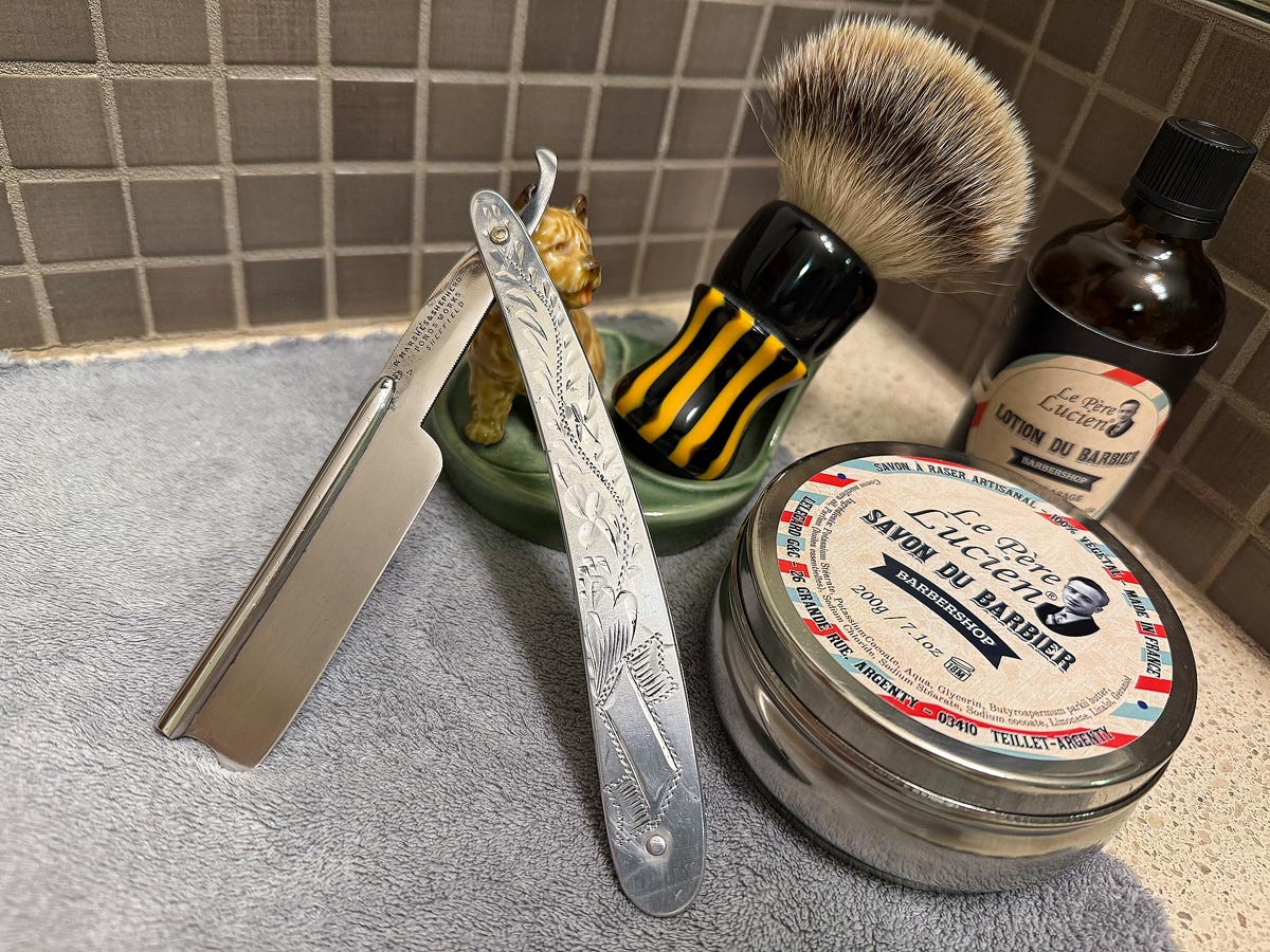 On a gray microfiber towel a straight razor with blade open about 45 degrees leans next to a bottle of aftershave and a jar of shave soap. A shave brush rests bristles-up in a green Wade ceramic pipe rest that features a brown Cairn Terrier.