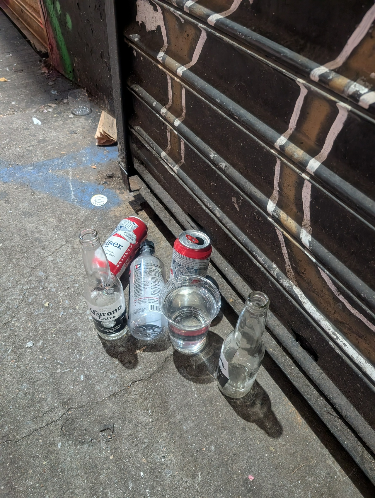 a group of liquid receptacles hangs around a closed shop gate: a few empty Coronas and Buds, one laying down along with a water bottle, and a completely full plastic cup of water in the middle of them all.