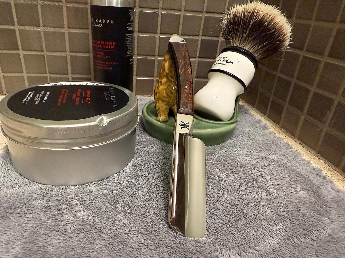 On a gray microfiber towel a kamisori style razor leans next to a bottle of aftershave and a jar of shave soap. A shave brush rests bristles-up in a green Wade ceramic pipe rest that features a brown Cairn Terrier.