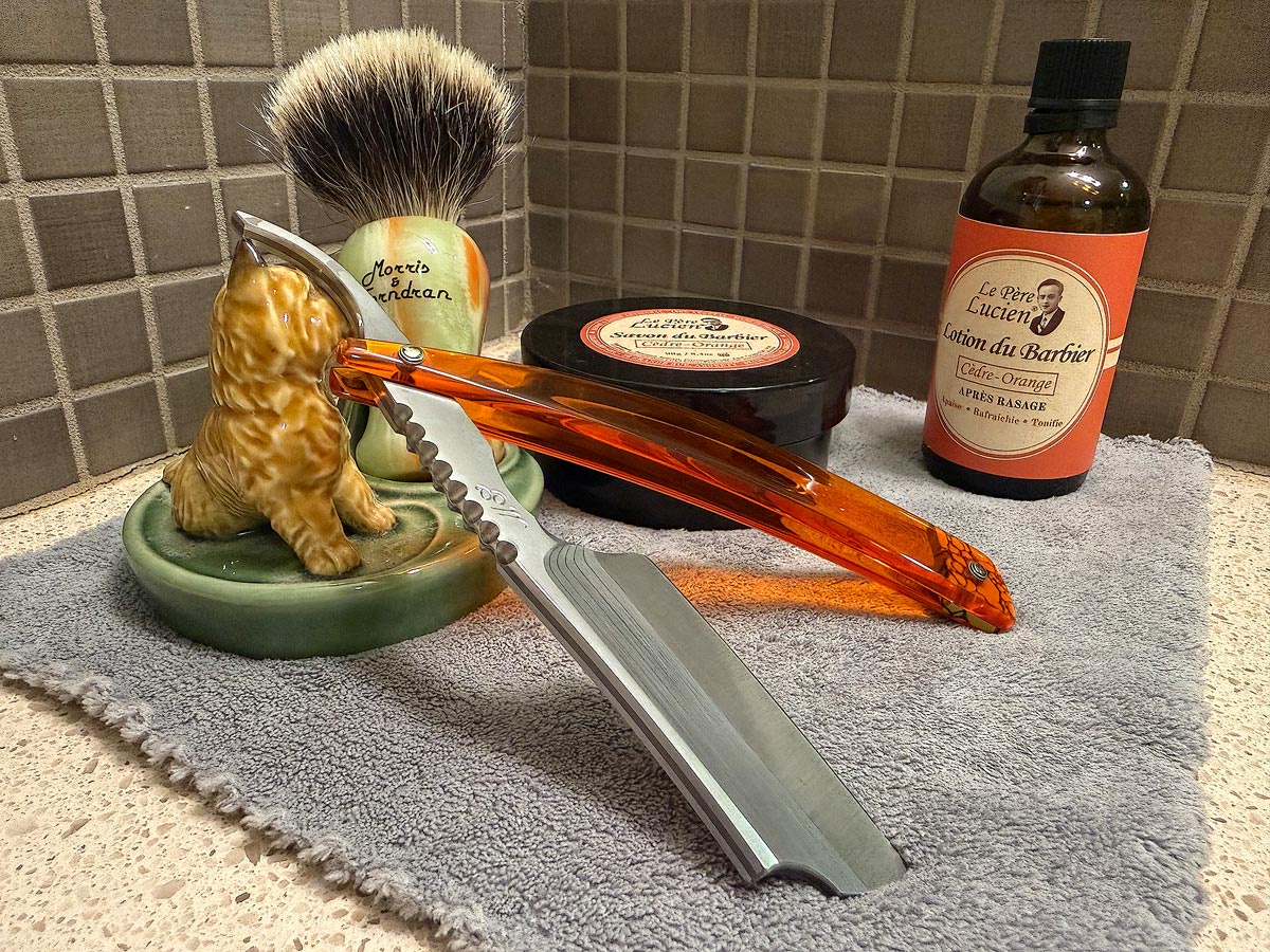 On a gray microfiber towel a straight razor with blade open about 45 degrees leans next to a bottle of aftershave and a jar of shave soap. A shave brush rests bristles-up in a green Wade ceramic pipe rest that features a brown Cairn Terrier.