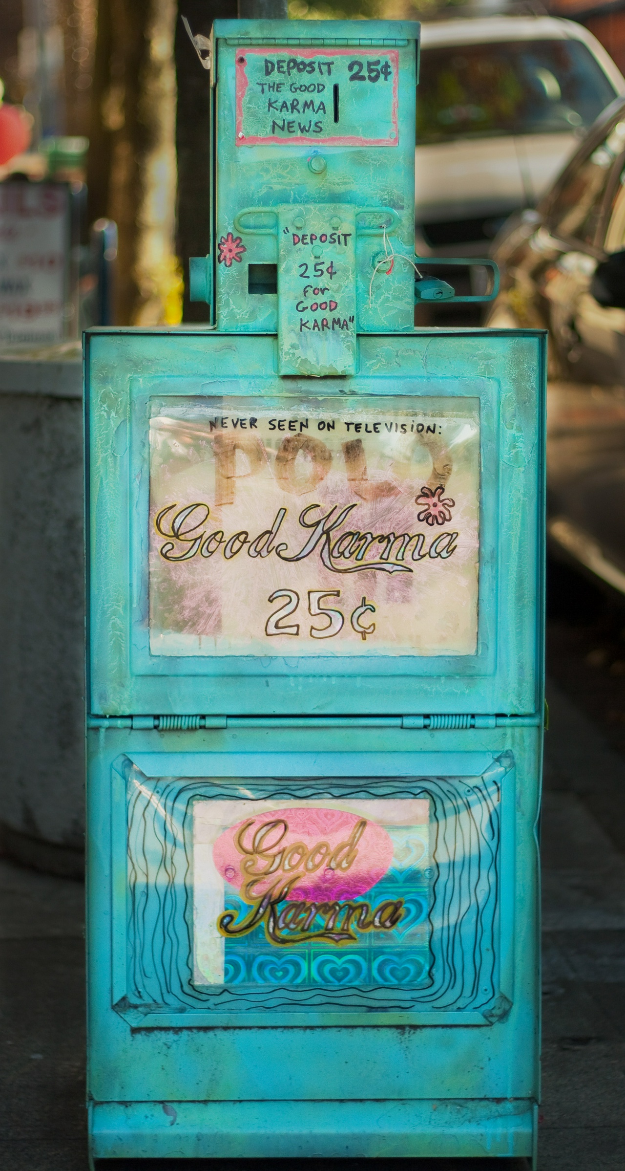 a brightly painted and slightly beat-up teal newspaper box, with pink accents, advertising Good Karma News - Deposit 25¢ for Good Karma!