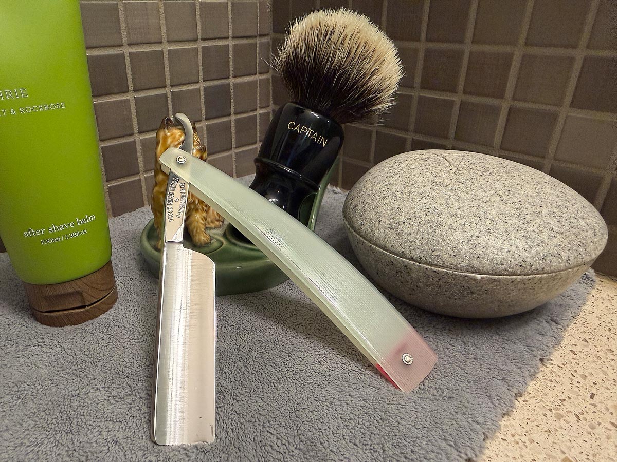 On a gray microfiber towel a straight razor with blade open about 45 degrees leans next to a bottle of aftershave and a jar of shave soap. A shave brush rests bristles-up in a green Wade ceramic pipe rest that features a brown Cairn Terrier.