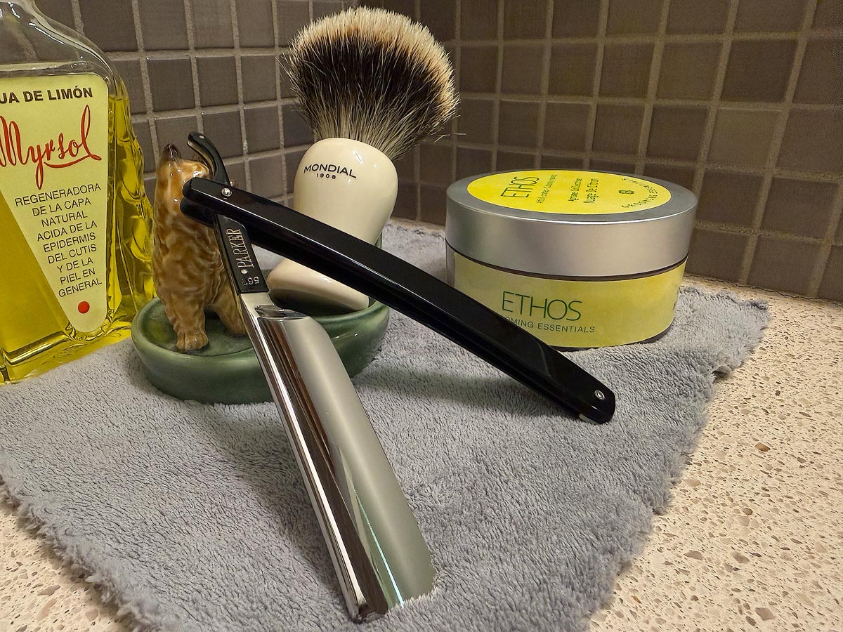 On a gray microfiber towel a straight razor with blade open about 45 degrees leans next to a bottle of aftershave and a jar of shave soap. A shave brush rests bristles-up in a green Wade ceramic pipe rest that features a brown Cairn Terrier.