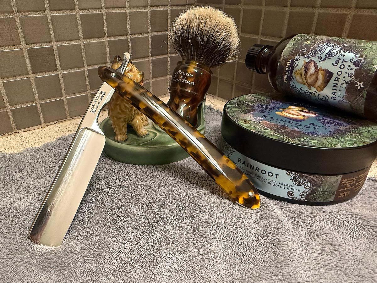 On a gray microfiber towel a straight razor with blade open about 45 degrees leans next to a bottle of aftershave and a jar of shave soap. A shave brush rests bristles-up in a green Wade ceramic pipe rest that features a brown Cairn Terrier.