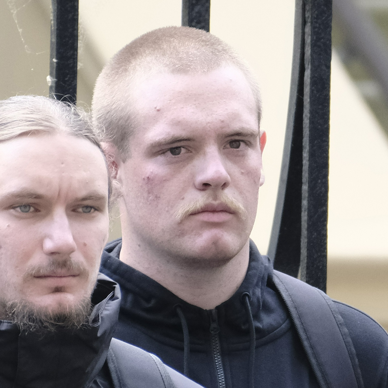 A pimply blonde man with a sparse moustache and a large round head wearing a black hoodie