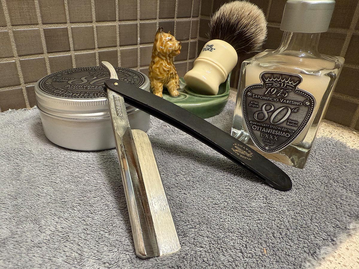 On a gray microfiber towel a straight razor with blade open about 45 degrees leans next to a bottle of aftershave and a jar of shave soap. A shave brush rests bristles-up in a green Wade ceramic pipe rest that features a brown Cairn Terrier.