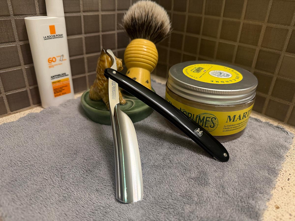 On a gray microfiber towel a straight razor with blade open about 45 degrees leans next to a bottle of aftershave and a jar of shave soap. A shave brush rests bristles-up in a green Wade ceramic pipe rest that features a brown Cairn Terrier.