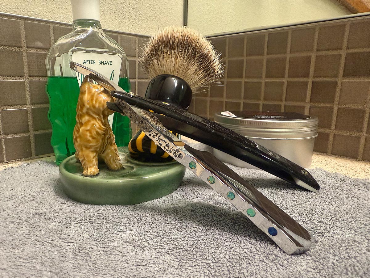 On a gray microfiber towel a straight razor with blade open about 45 degrees leans next to a bottle of aftershave and a jar of shave soap. A shave brush rests bristles-up in a green Wade ceramic pipe rest that features a brown Cairn Terrier.