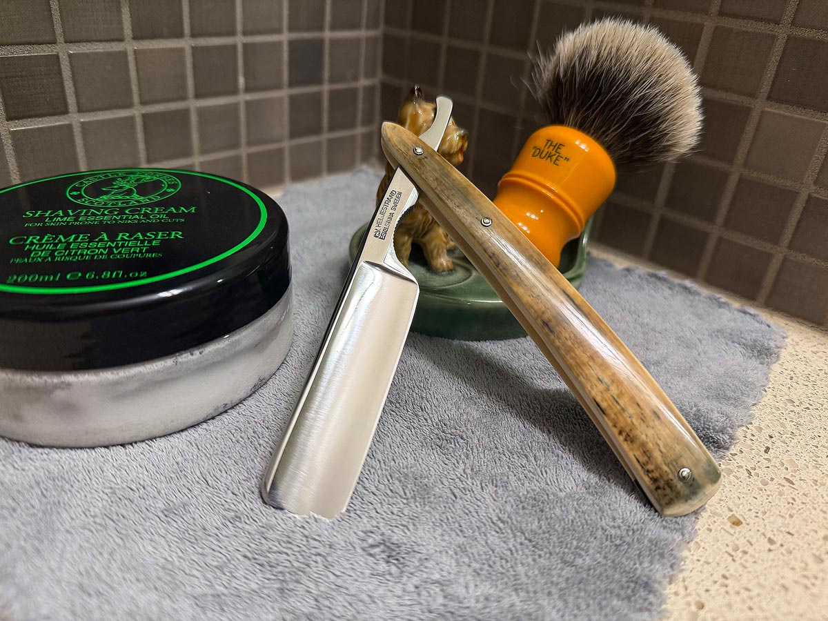 On a gray microfiber towel a straight razor with blade open about 45 degrees leans next to a bottle of aftershave and a jar of shave soap. A shave brush rests bristles-up in a green Wade ceramic pipe rest that features a brown Cairn Terrier.