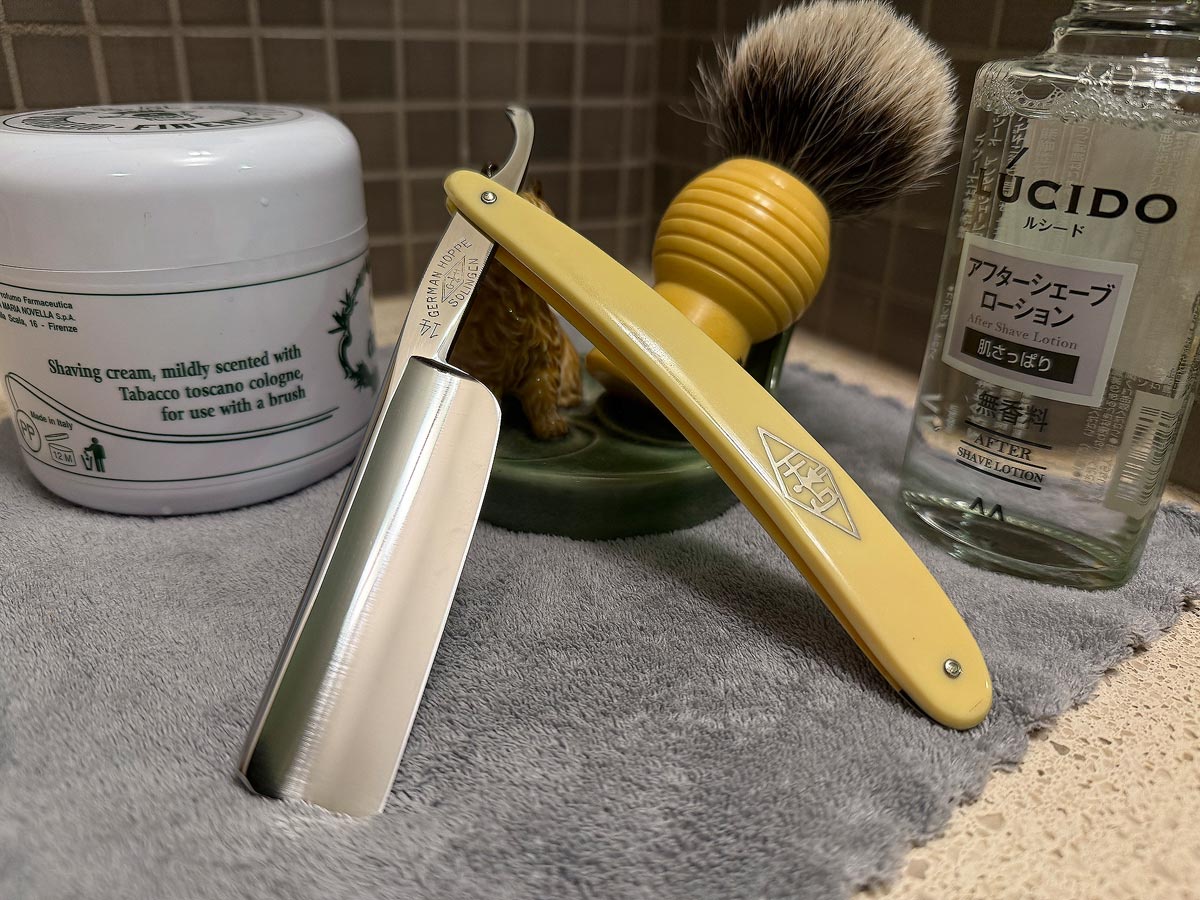 On a gray microfiber towel a straight razor with blade open about 45 degrees leans next to a bottle of aftershave and a jar of shave soap. A shave brush rests bristles-up in a green Wade ceramic pipe rest that features a brown Cairn Terrier.