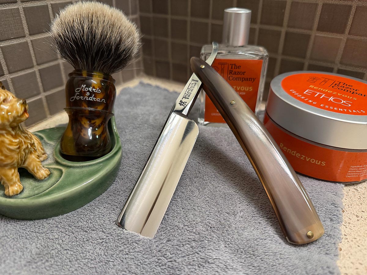 On a gray microfiber towel a straight razor with blade open about 45 degrees leans next to a bottle of aftershave and a jar of shave soap. A shave brush rests bristles-up in a green Wade ceramic pipe rest that features a brown Cairn Terrier.
