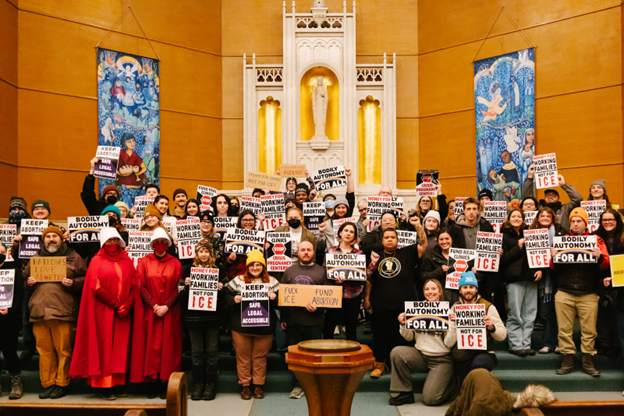 Activists gather for a group photo in a church, holding signs with slogans including “Bodily autonomy for all,” “Keep abortion safe, legal, accessible” and “Money for working families, not for ICE.” | Watch Me Rise Minneapolis Activists gather for a group photo in a church, holding signs with slogans including “Bodily autonomy for all,” “Keep abortion safe, legal, accessible” and “Money for working families, not for ICE.”