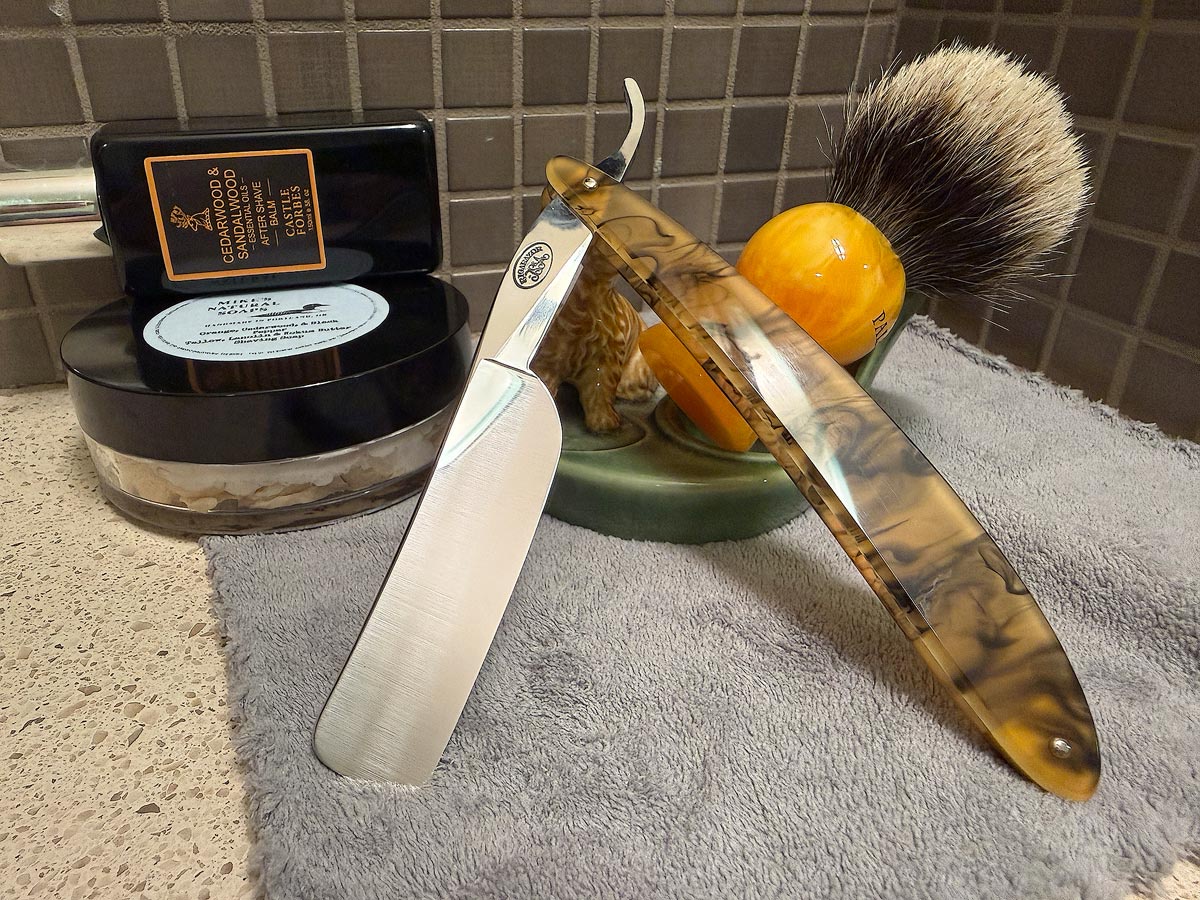 On a gray microfiber towel a straight razor with blade open about 45 degrees leans next to a bottle of aftershave and a jar of shave soap. A shave brush rests bristles-up in a green Wade ceramic pipe rest that features a brown Cairn Terrier.