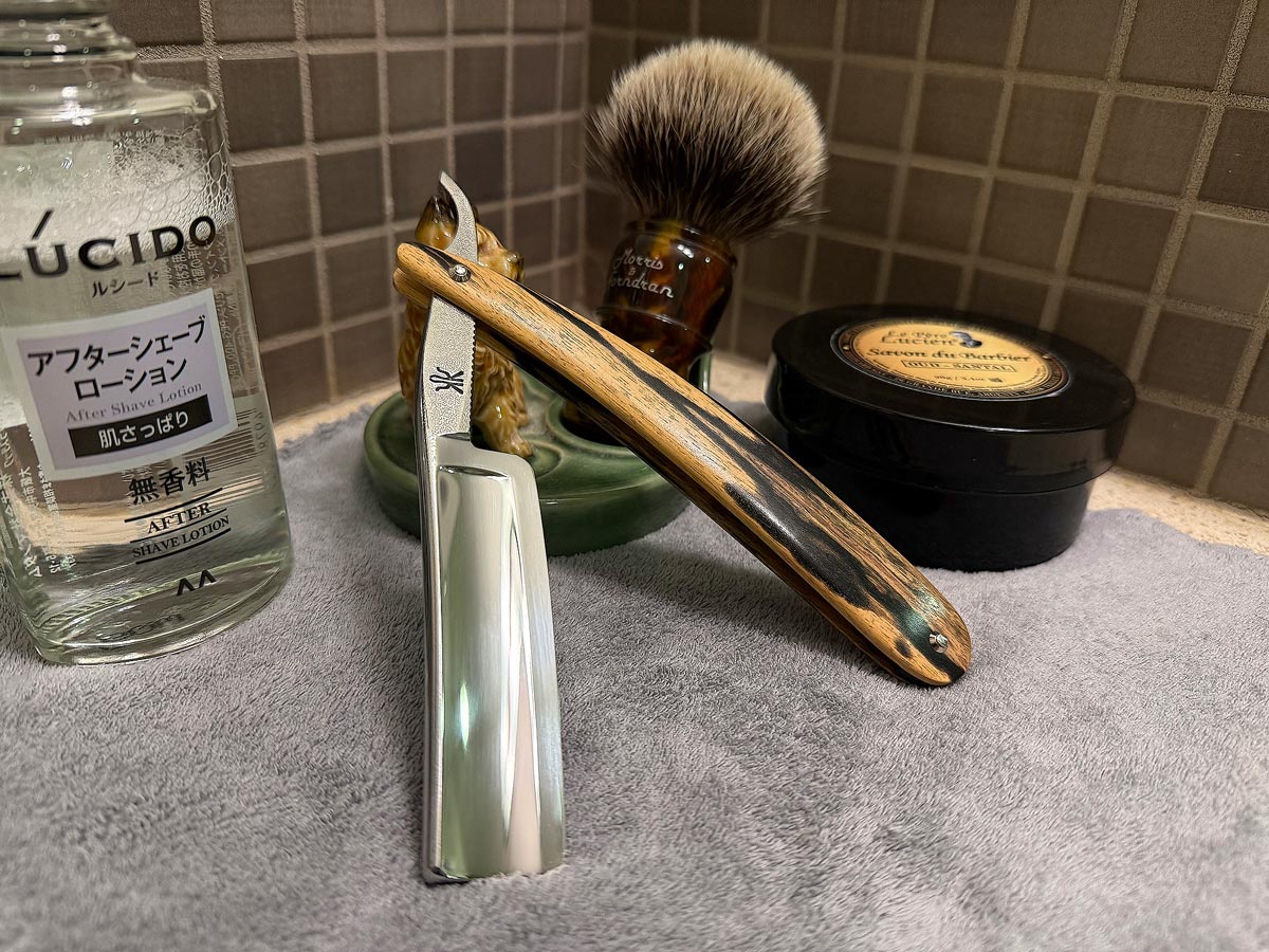 On a gray microfiber towel a straight razor with blade open about 45 degrees leans next to a bottle of aftershave and a jar of shave soap. A shave brush rests bristles-up in a green Wade ceramic pipe rest that features a brown Cairn Terrier.