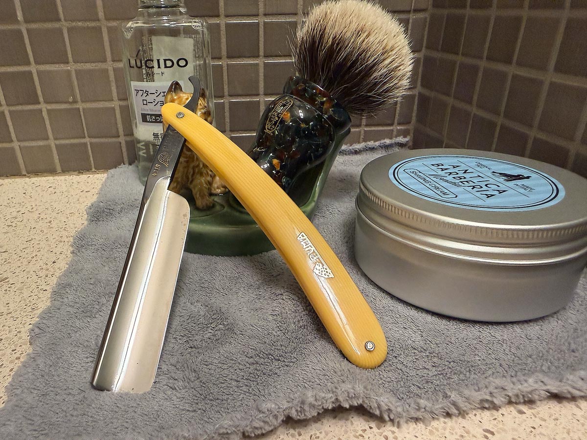 On a gray microfiber towel a straight razor with blade open about 45 degrees leans next to a bottle of aftershave and a jar of shave soap. A shave brush rests bristles-up in a green Wade ceramic pipe rest that features a brown Cairn Terrier.