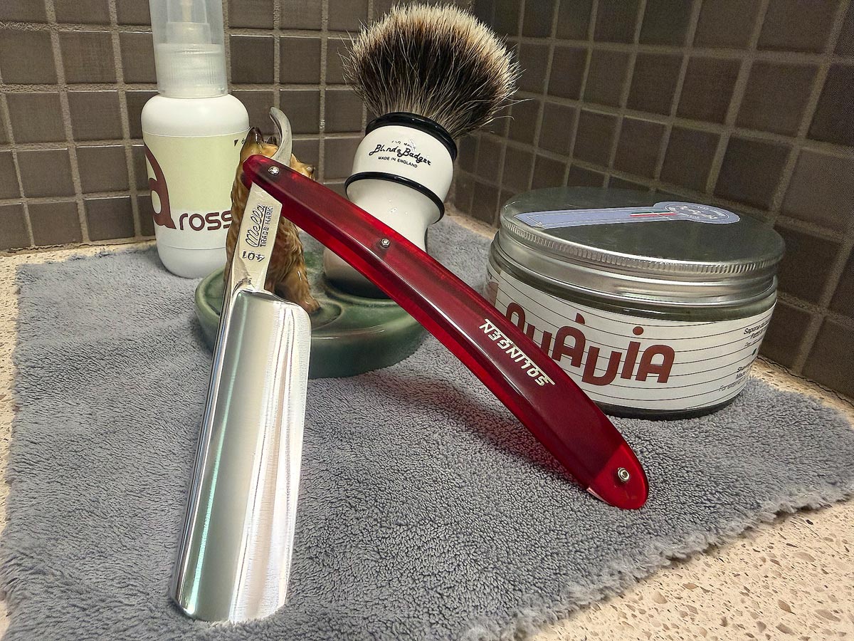 On a gray microfiber towel a straight razor with blade open about 45 degrees leans next to a bottle of aftershave and a jar of shave soap. A shave brush rests bristles-up in a green Wade ceramic pipe rest that features a brown Cairn Terrier.