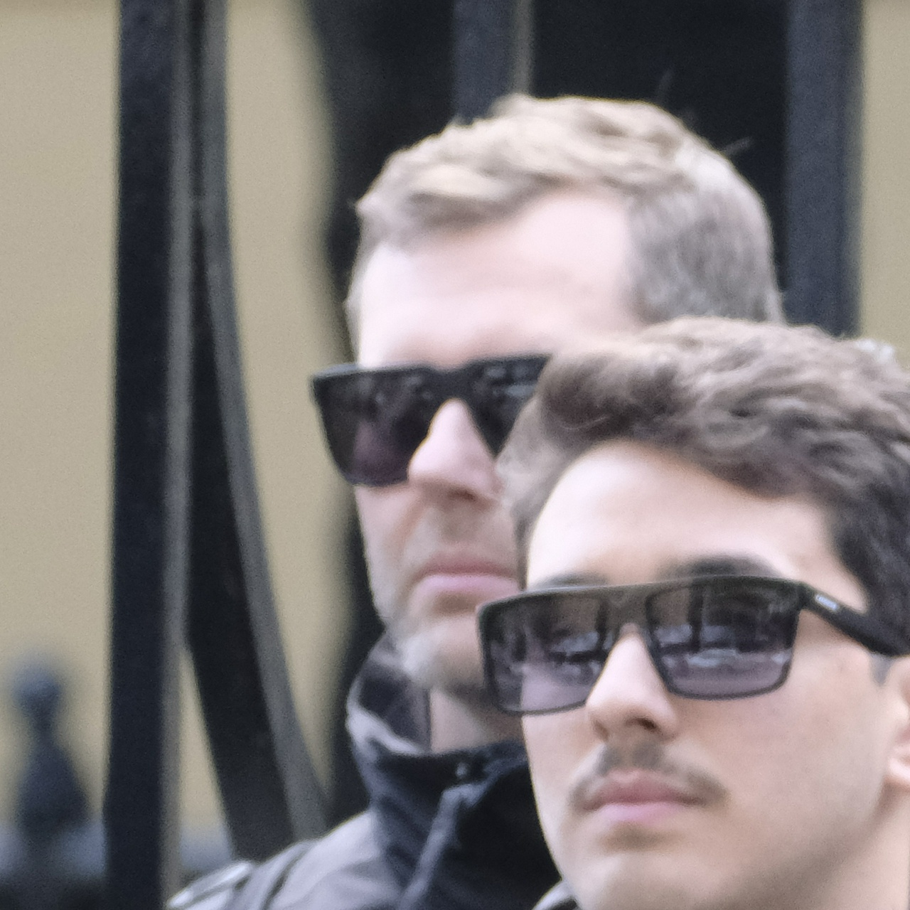 A tall older guy with short, light coloured hair peeks out from behind his companion, frowning