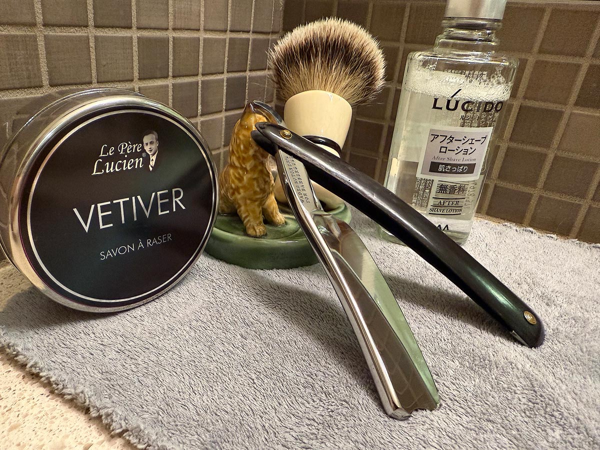 On a gray microfiber towel a straight razor with blade open about 45 degrees leans next to a bottle of aftershave and a jar of shave soap. A shave brush rests bristles-up in a green Wade ceramic pipe rest that features a brown Cairn Terrier.
