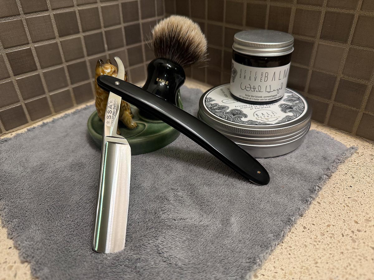 On a gray microfiber towel a straight razor with blade open about 45 degrees leans next to a bottle of aftershave and a jar of shave soap. A shave brush rests bristles-up in a green Wade ceramic pipe rest that features a brown Cairn Terrier.