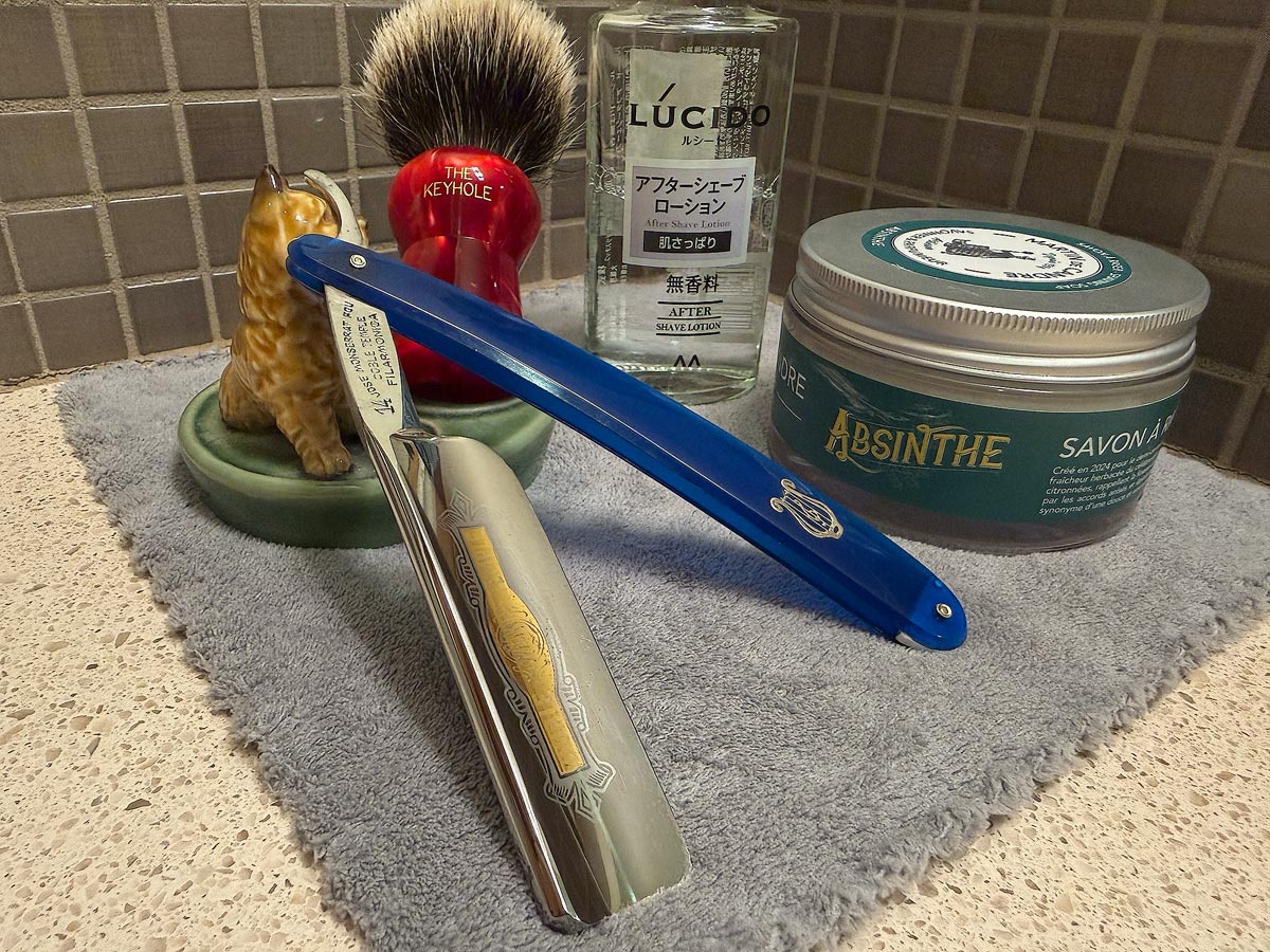 On a gray microfiber towel a straight razor with blade open about 45 degrees leans next to a bottle of aftershave and a jar of shave soap. A shave brush rests bristles-up in a green Wade ceramic pipe rest that features a brown Cairn Terrier.