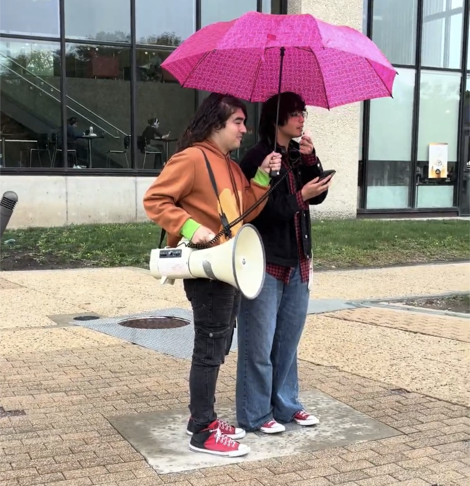 Two student protesters stand holding a megaphone and an umbrella.