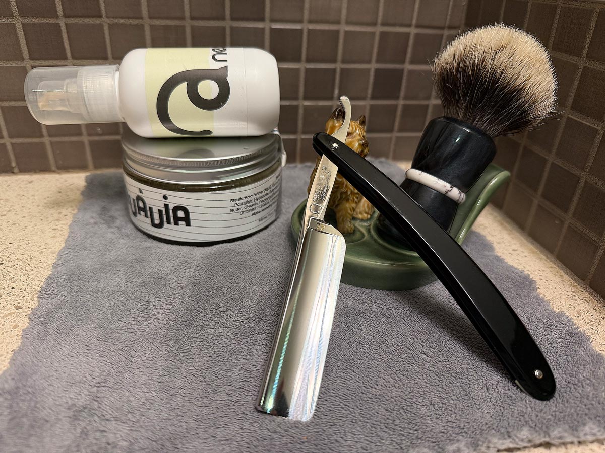 On a gray microfiber towel a straight razor with blade open about 45 degrees leans next to a bottle of aftershave and a jar of shave soap. A shave brush rests bristles-up in a green Wade ceramic pipe rest that features a brown Cairn Terrier.