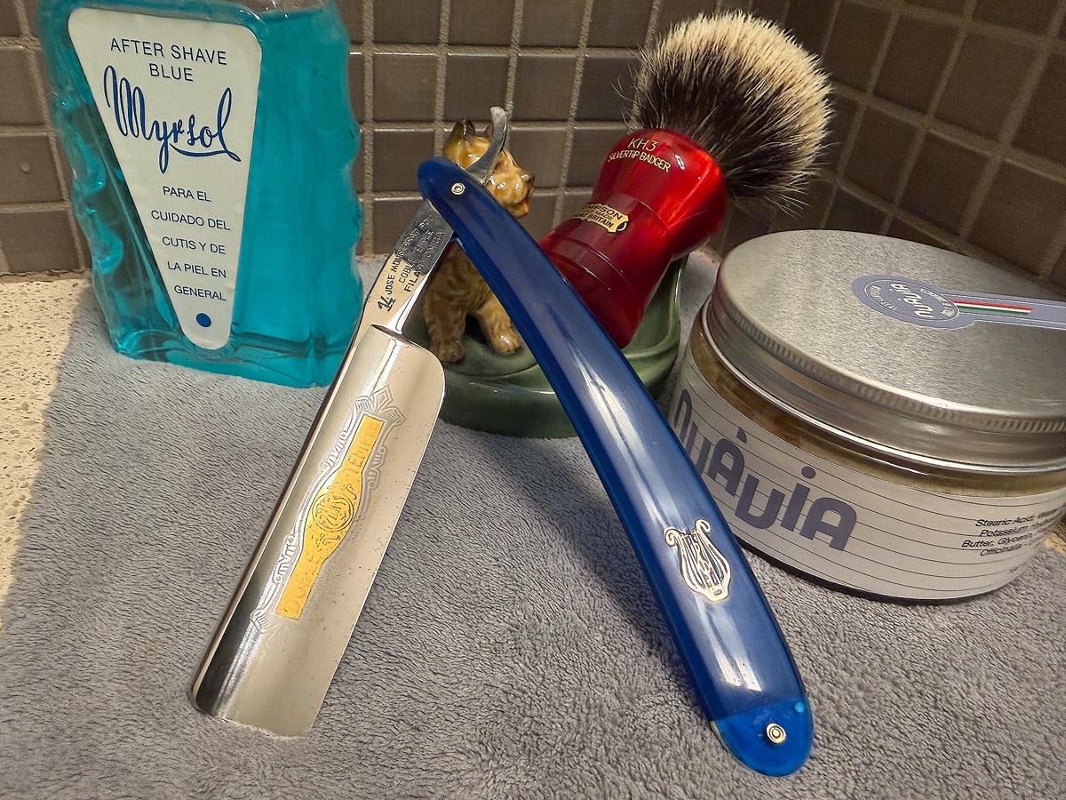 On a gray microfiber towel a straight razor with blade open about 45 degrees leans next to a bottle of aftershave and a jar of shave soap. A shave brush rests bristles-up in a green Wade ceramic pipe rest that features a brown Cairn Terrier.