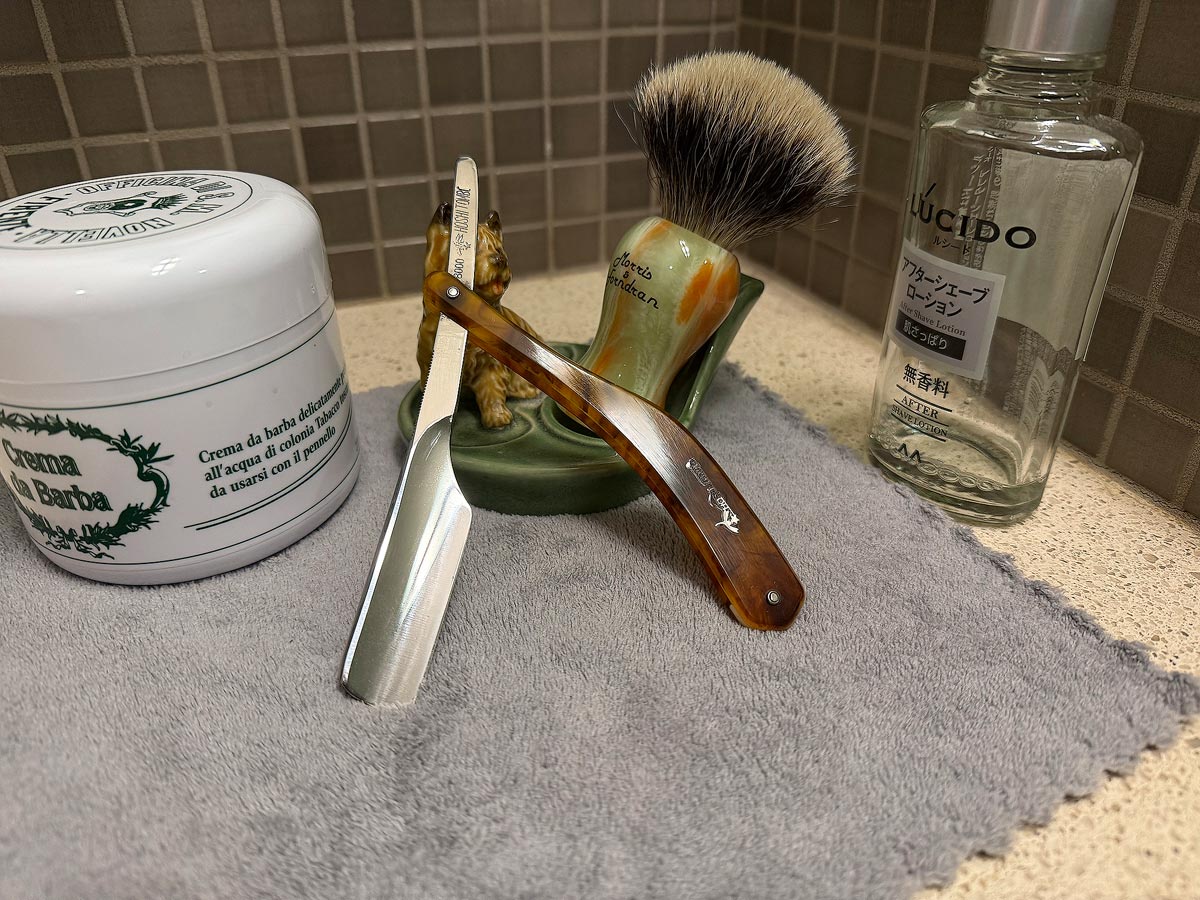 On a gray microfiber towel a straight razor with blade open about 45 degrees leans next to a bottle of aftershave and a jar of shave soap. A shave brush rests bristles-up in a green Wade ceramic pipe rest that features a brown Cairn Terrier.
