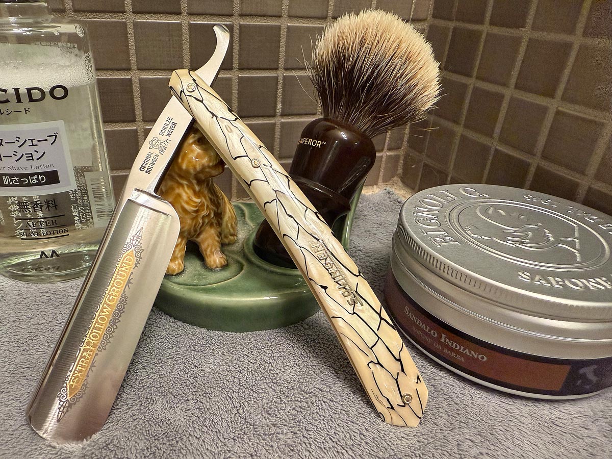 On a gray microfiber towel a straight razor with blade open about 45 degrees leans next to a bottle of aftershave and a jar of shave soap. A shave brush rests bristles-up in a green Wade ceramic pipe rest that features a brown Cairn Terrier.