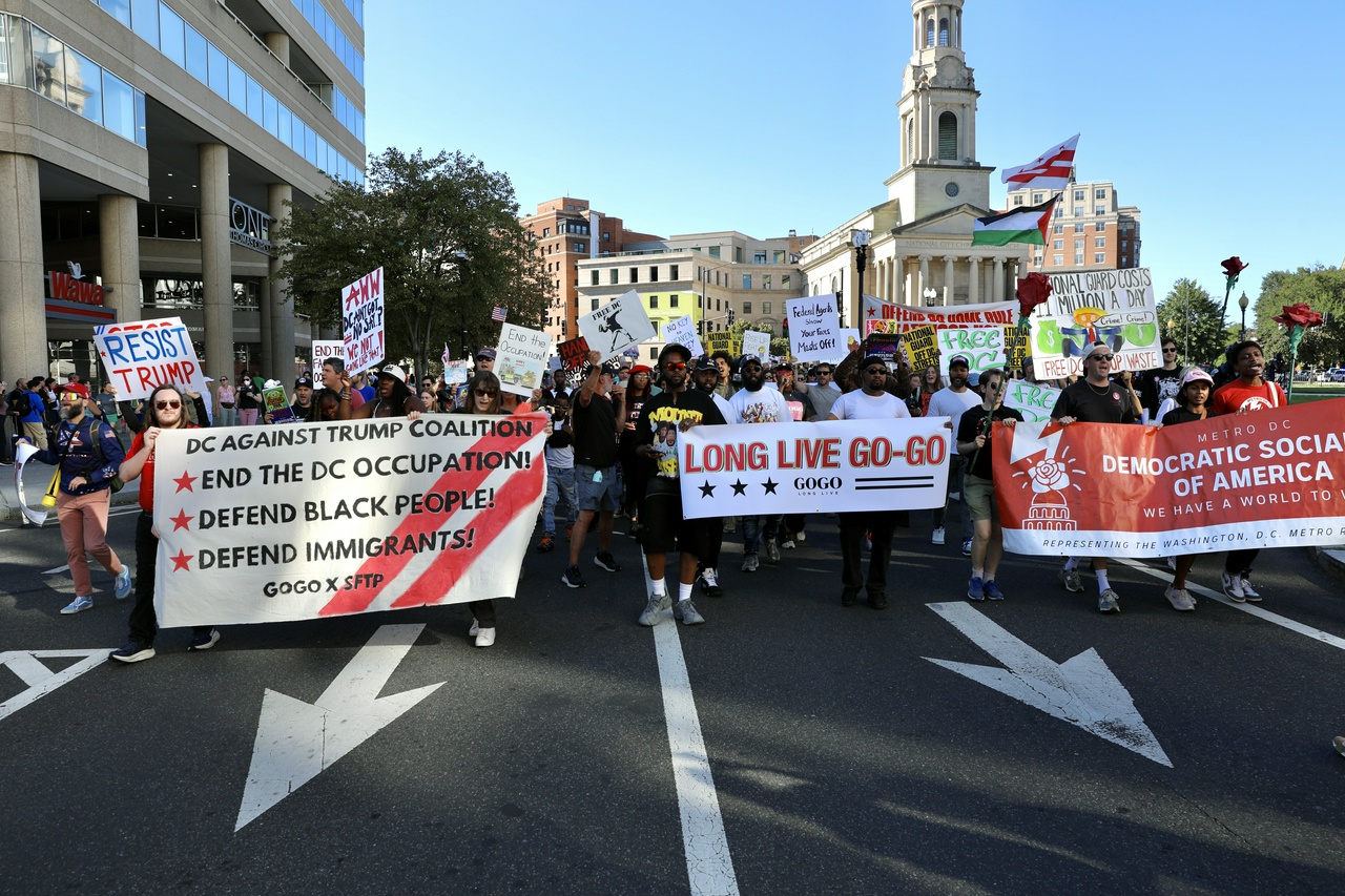 Protesters march through DC with the White House capitol building in the background. They hold banenrs saying "End the DC Occupation! Defend Black People! Defend Immigrants!"