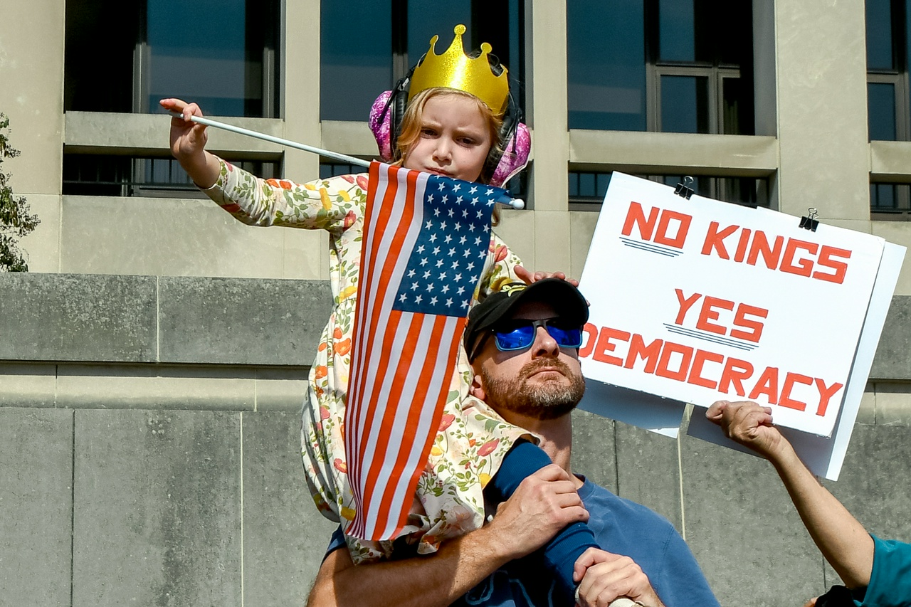 Man with daughter wearing a crown sitting on his shoulder, next to protest sign saying No Kings, Yes Democracy.