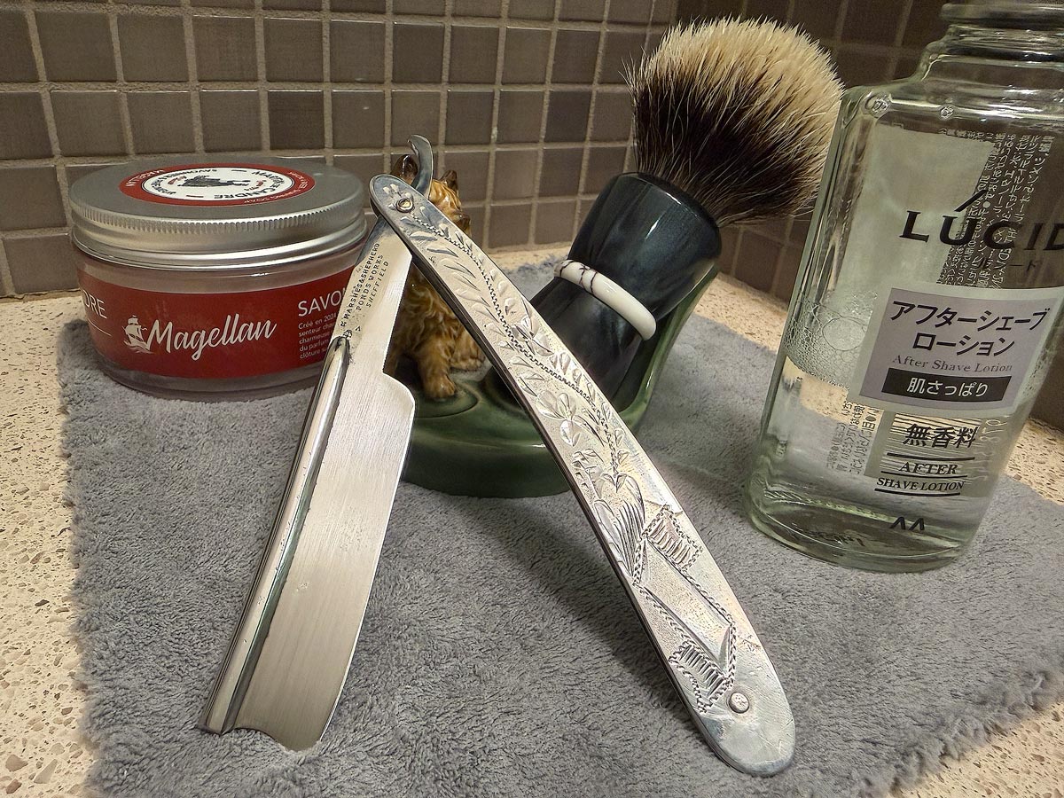 On a gray microfiber towel a straight razor with blade open about 45 degrees leans next to a bottle of aftershave and a jar of shave soap. A shave brush rests bristles-up in a green Wade ceramic pipe rest that features a brown Cairn Terrier.
