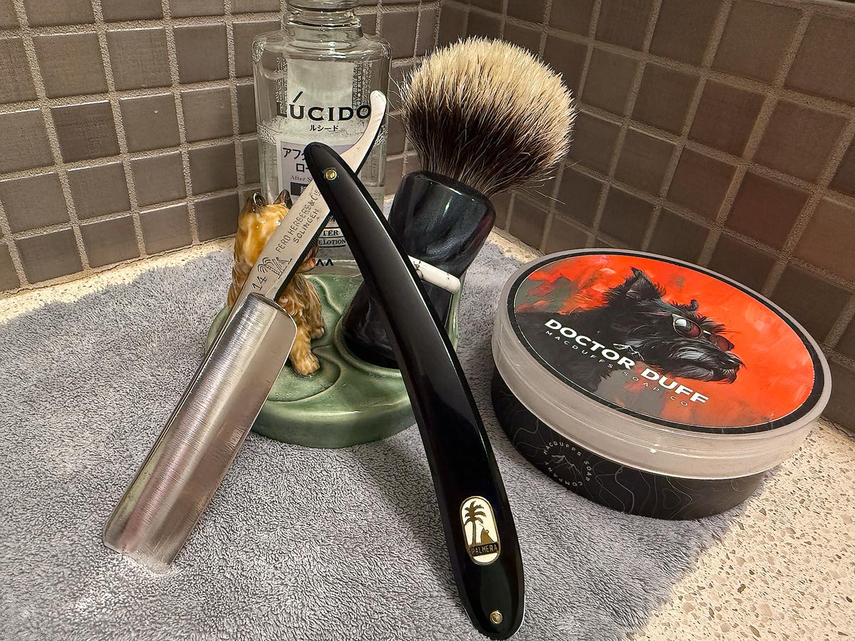 On a gray microfiber towel a straight razor with blade open about 45 degrees leans next to a bottle of aftershave and a jar of shave soap. A shave brush rests bristles-up in a green Wade ceramic pipe rest that features a brown Cairn Terrier.