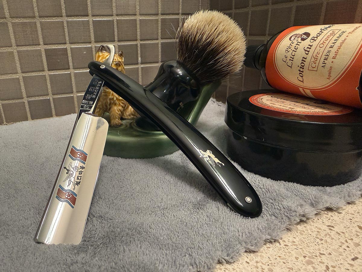 On a gray microfiber towel a straight razor with blade open about 45 degrees leans next to a bottle of aftershave and a jar of shave soap. A shave brush rests bristles-up in a green Wade ceramic pipe rest that features a brown Cairn Terrier.
