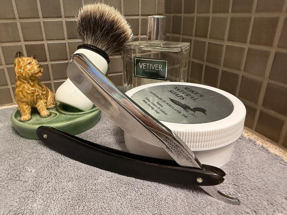 On a gray microfiber towel a straight razor with blade open about 45 degrees leans next to a bottle of aftershave and a jar of shave soap. A shave brush rests bristles-up in a green Wade ceramic pipe rest that features a brown Cairn Terrier.