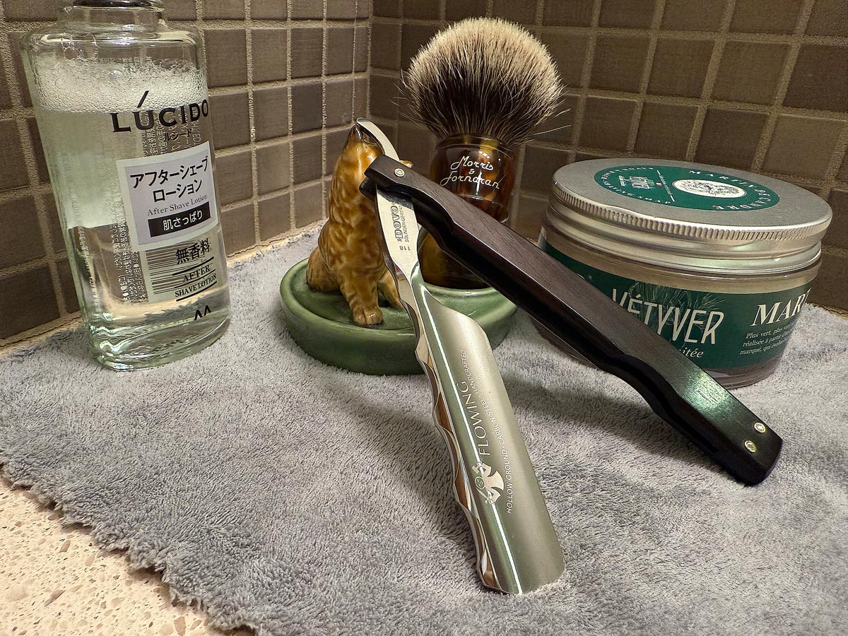 On a gray microfiber towel a straight razor with blade open about 45 degrees leans next to a bottle of aftershave and a jar of shave soap. A shave brush rests bristles-up in a green Wade ceramic pipe rest that features a brown Cairn Terrier.