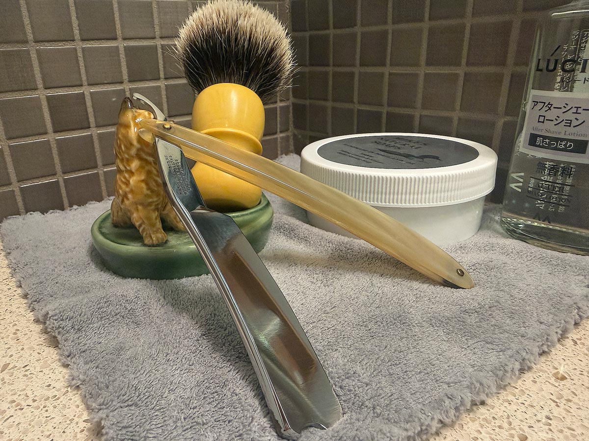 On a gray microfiber towel a straight razor with blade open about 45 degrees leans next to a bottle of aftershave and a jar of shave soap. A shave brush rests bristles-up in a green Wade ceramic pipe rest that features a brown Cairn Terrier.