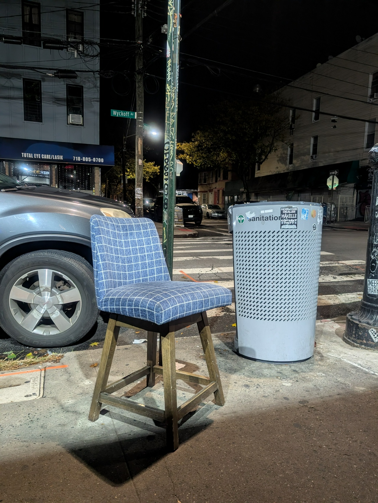 a very handsome, cushy chair with a blue and white grid and wooden legs is comfortably perched near the curb in brooklyn, against a street sign post covered in graffiti, near a city trashcan