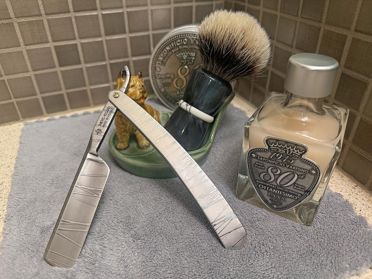 On a gray microfiber towel a straight razor with blade open about 45 degrees leans next to a bottle of aftershave and a jar of shave soap. A shave brush rests bristles-up in a green Wade ceramic pipe rest that features a brown Cairn Terrier.