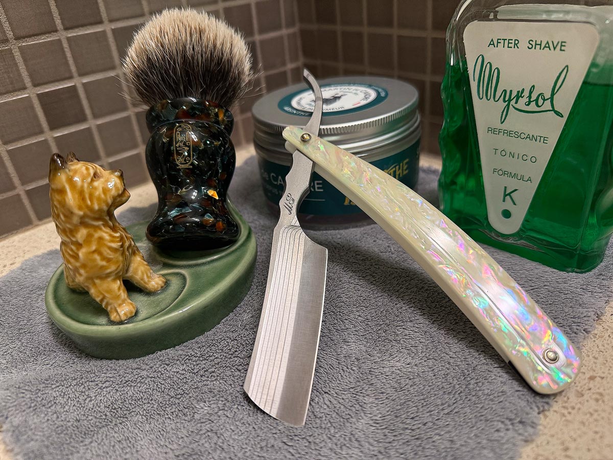 On a gray microfiber towel a straight razor with blade open about 45 degrees leans next to a bottle of aftershave and a jar of shave soap. A shave brush rests bristles-up in a green Wade ceramic pipe rest that features a brown Cairn Terrier.