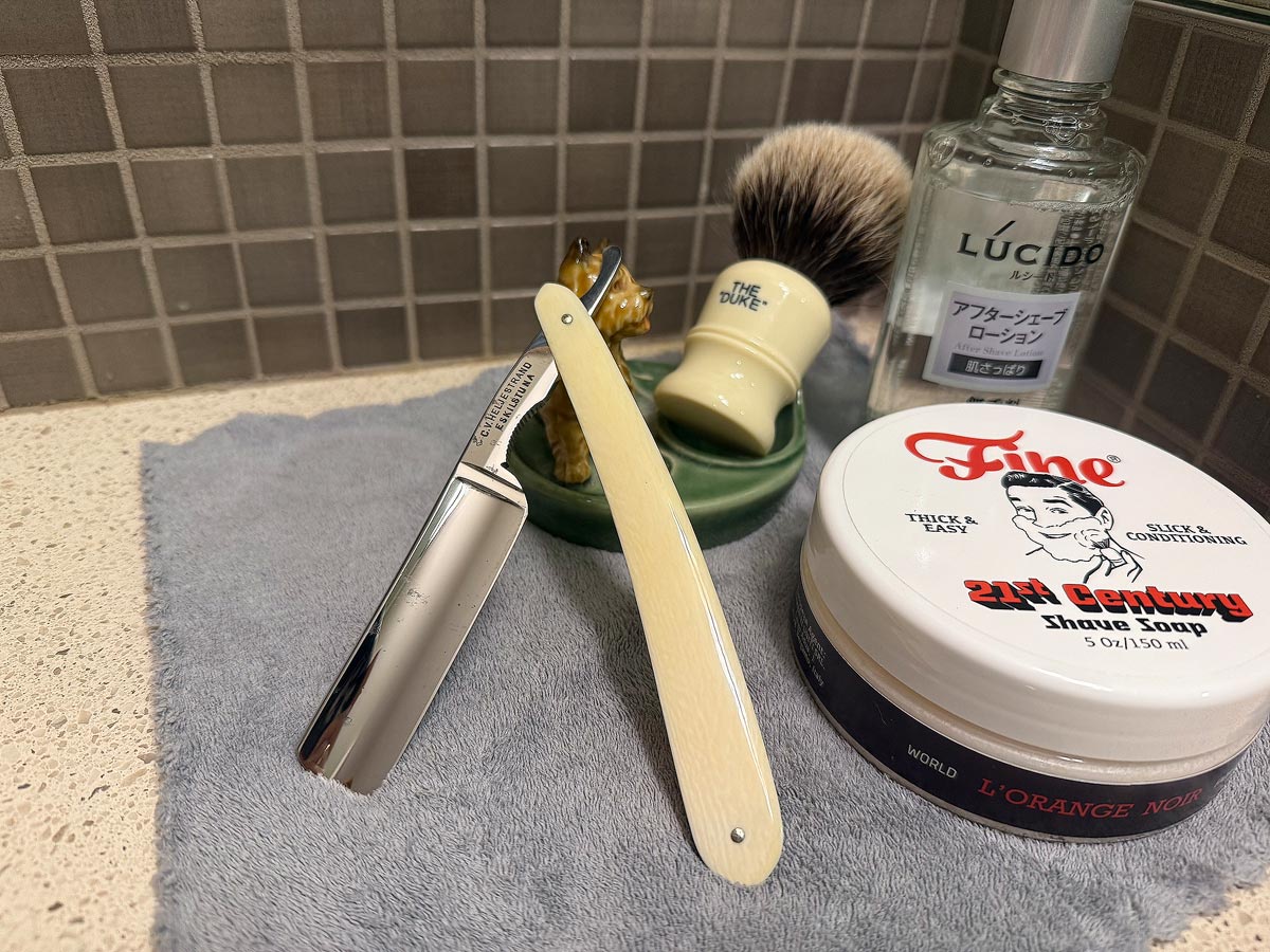 On a gray microfiber towel a straight razor with blade open about 45 degrees leans next to a bottle of aftershave and a jar of shave soap. A shave brush rests bristles-up in a green Wade ceramic pipe rest that features a brown Cairn Terrier.