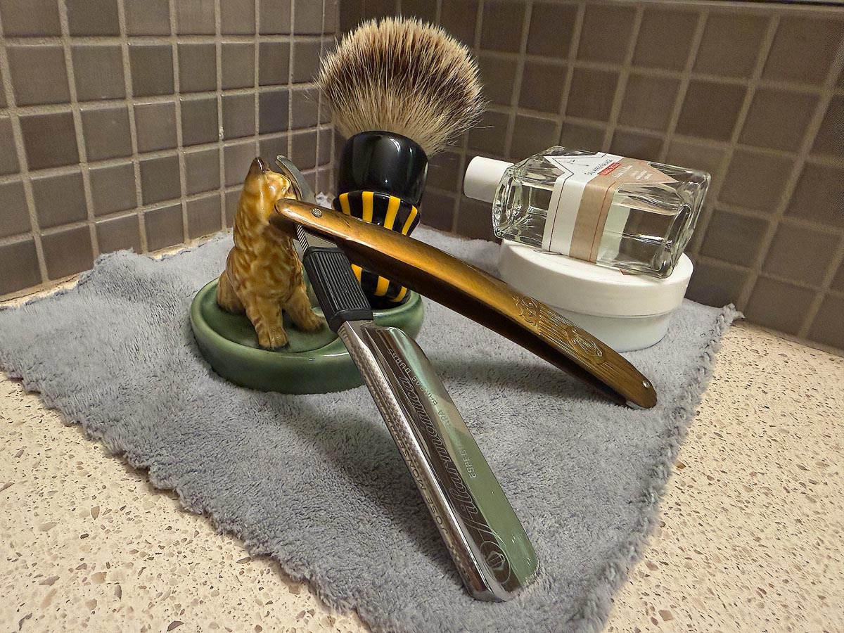 On a gray microfiber towel a straight razor with blade open about 45 degrees leans next to a bottle of aftershave and a jar of shave soap. A shave brush rests bristles-up in a green Wade ceramic pipe rest that features a brown Cairn Terrier.
