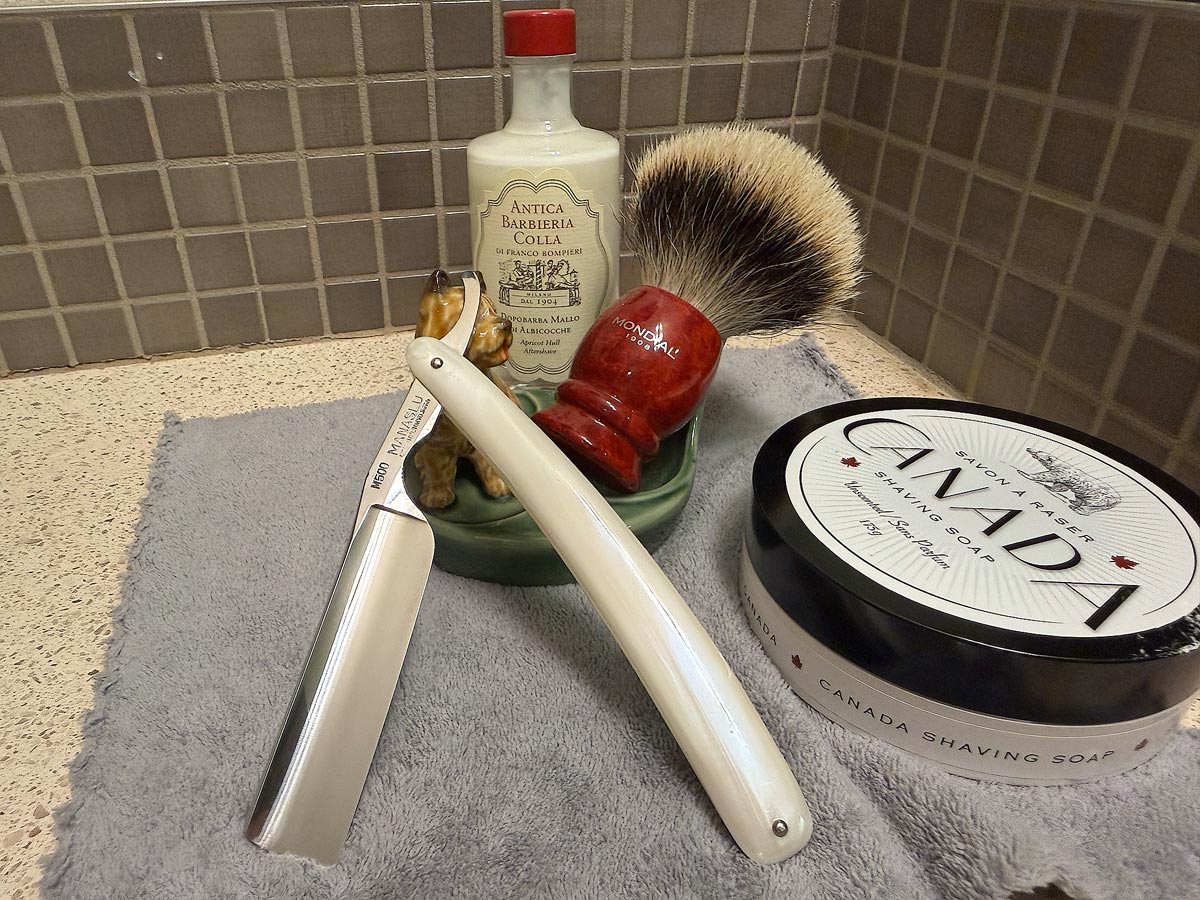 On a gray microfiber towel a straight razor with blade open about 45 degrees leans next to a bottle of aftershave and a jar of shave soap. A shave brush rests bristles-up in a green Wade ceramic pipe rest that features a brown Cairn Terrier.