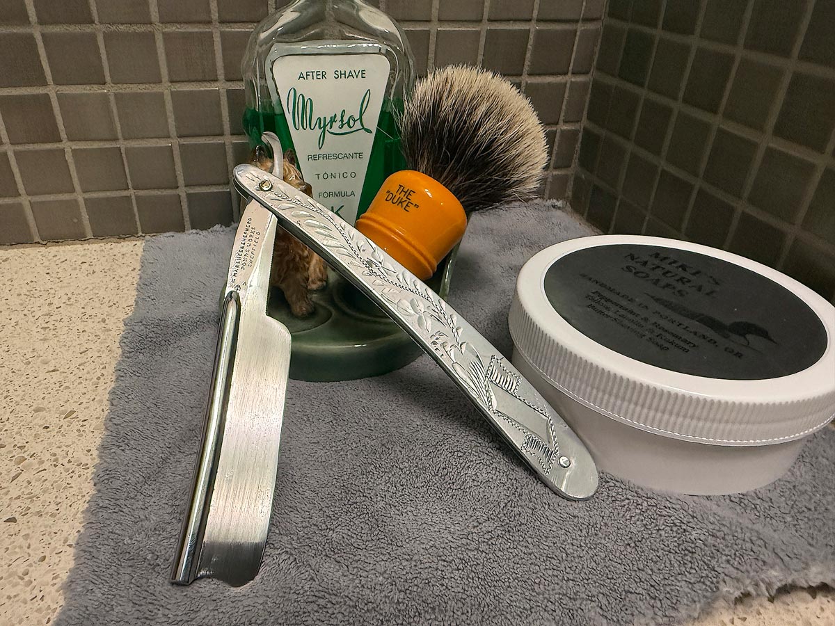 On a gray microfiber towel a straight razor with blade open about 45 degrees leans next to a bottle of aftershave and a jar of shave soap. A shave brush rests bristles-up in a green Wade ceramic pipe rest that features a brown Cairn Terrier.