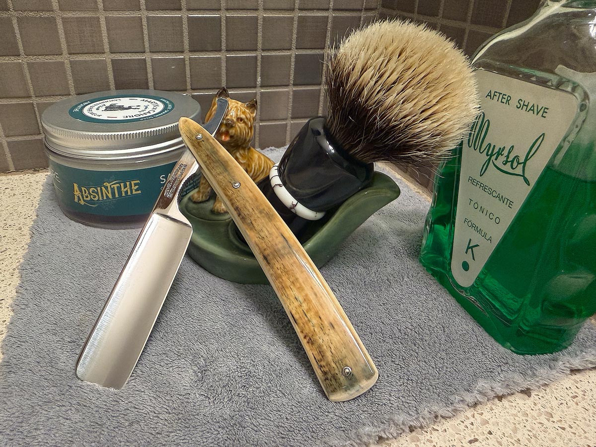 On a gray microfiber towel a straight razor with blade open about 45 degrees leans next to a bottle of aftershave and a jar of shave soap. A shave brush rests bristles-up in a green Wade ceramic pipe rest that features a brown Cairn Terrier.